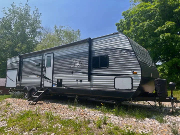 This RV rental is parked on a gravel lot surrounded by green trees under a clear blue sky, ready for your next adventure.. Jayco Jay Flight 2021