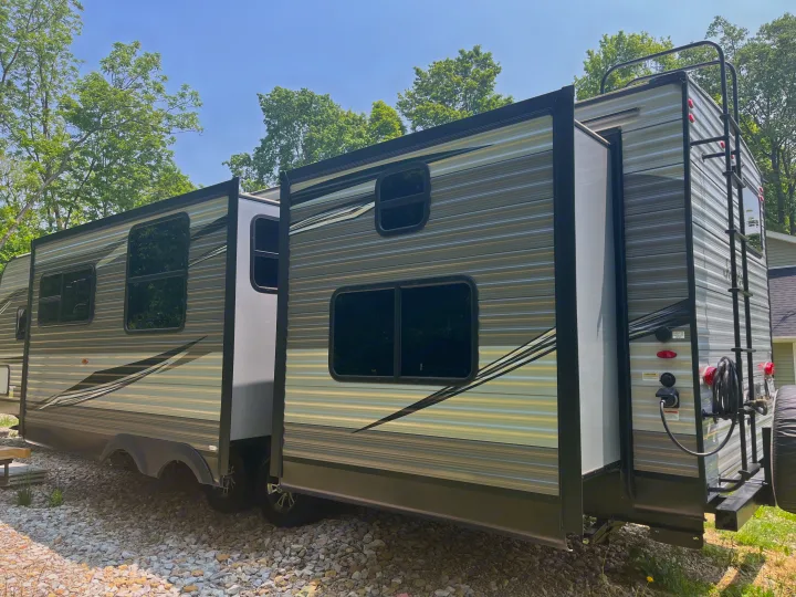 Side view of an RV rental: a modern travel trailer with slide-outs, windows, and rear ladder, on gravel.. Jayco Jay Flight 2021