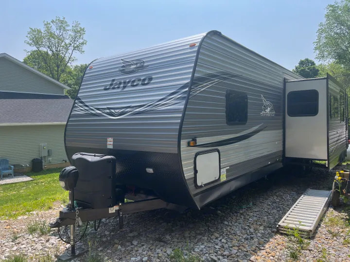 A modern grey Jayco travel trailer, ideal as an RV rental, is parked on gravel beside a light green house.. Jayco Jay Flight 2021