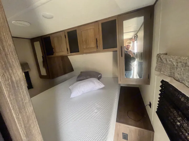 Inside an RV rental: a compact bedroom with a white mattress, overhead wood cabinets, and a side mirror.. Avenger Avenger Trailer 2021