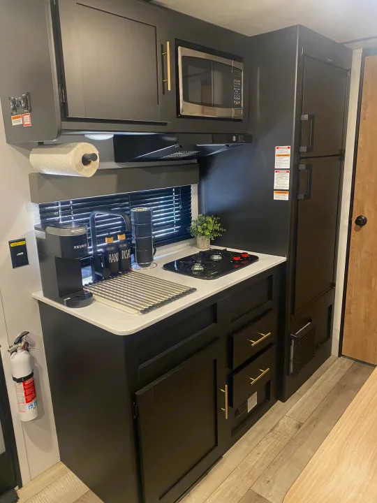 Compact, modern RV rental kitchen with dark cabinets. Features a microwave, stovetop, sink, and coffee maker, ideal for travel.. Keystone RV Springdale 2022