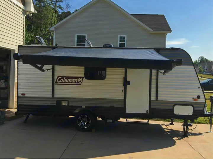 A beige and grey Coleman travel trailer is parked on a driveway with its black awning extended. Ideal for an rv rental.. Dutchmen Coleman Lantern LT 2020