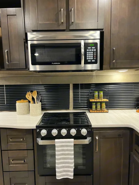 Modern RV rental kitchen featuring a Furrion microwave, gas oven, dark cabinets, and white countertops.. Grand Design Reflection 2021
