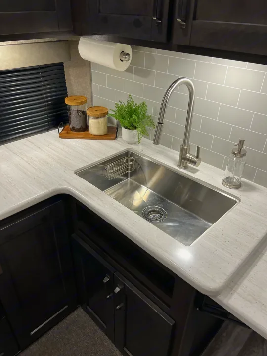 Modern RV rental kitchen: sleek stainless sink, gray tile backsplash, under-cabinet lighting, and counter items.. Grand Design Reflection 2021