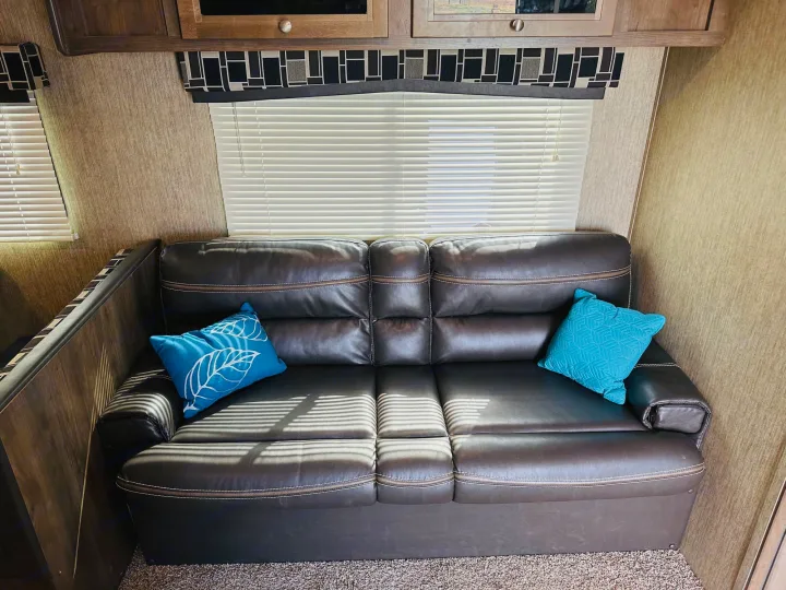 RV rental living area: a brown sofa with blue pillows, bright windows with blinds, and overhead cabinets.. Dutchmen Coleman Lantern 2019