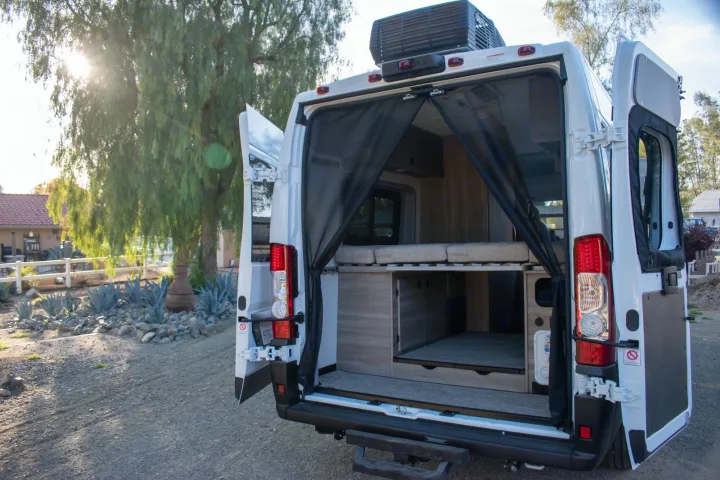 Open rear doors of a white camper van RV rental, showing its mesh screen, raised bed, and practical storage space.. Winnebago Solis 59PX 2023