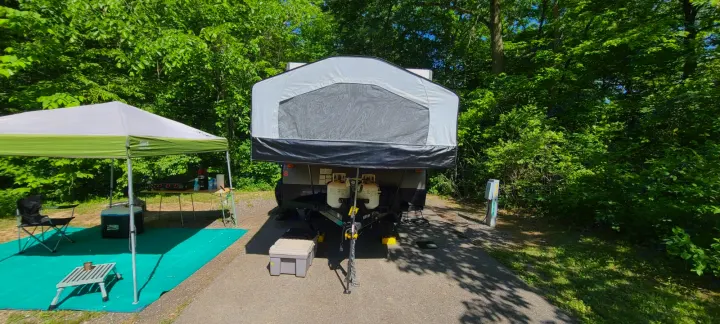 Pop-up RV rental set up at a sunny campsite with a shaded canopy, outdoor kitchen, and power hookup amidst green trees.. Forest River Rockwood ESP 2017