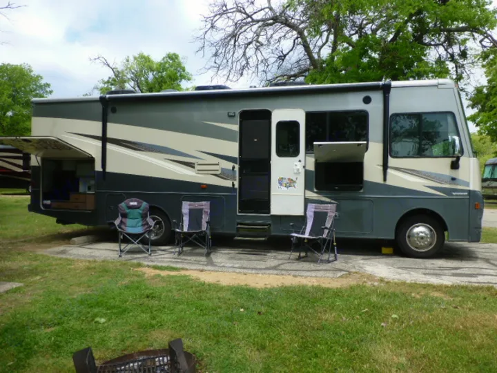 Spacious RV rental motorhome at a shaded campsite. Chairs and open bays create an inviting outdoor living area.. Winnebago Vista 2020