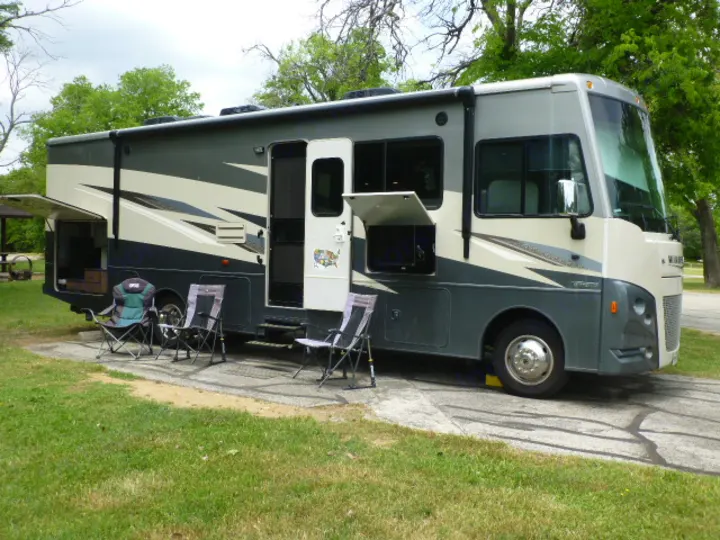 This grey and beige rv rental is set up at a campground, with open compartments and chairs for outdoor relaxation.. Winnebago Vista 2020