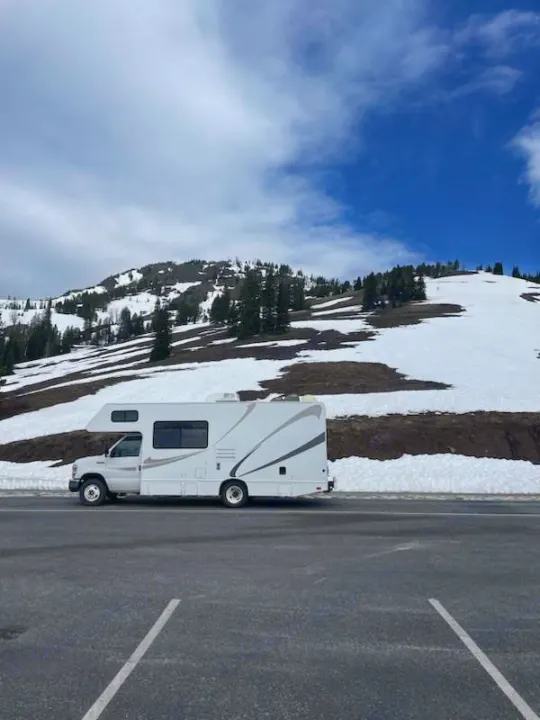 A white RV rental is parked on an asphalt lot beside a snow-covered mountain with scattered trees and blue sky.. Thor Majestic 2017