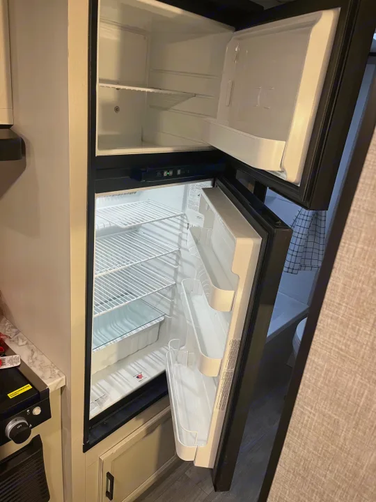 Inside an RV rental, the empty two-door refrigerator and freezer unit is open, revealing white shelves and bins.. Jayco Jay Flight SLX 2023