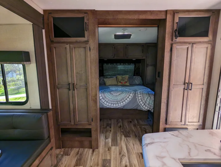 View inside an RV rental, highlighting the bedroom with decorative bedding and the adjacent living area's wood-look interior.. Sport Trek Sport Trek Trailer 2021