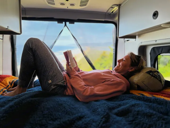 A woman smiles while relaxing and reading a book inside a cozy RV rental, with a scenic outdoor view visible through the rear window.. Winnebago Solis 2023