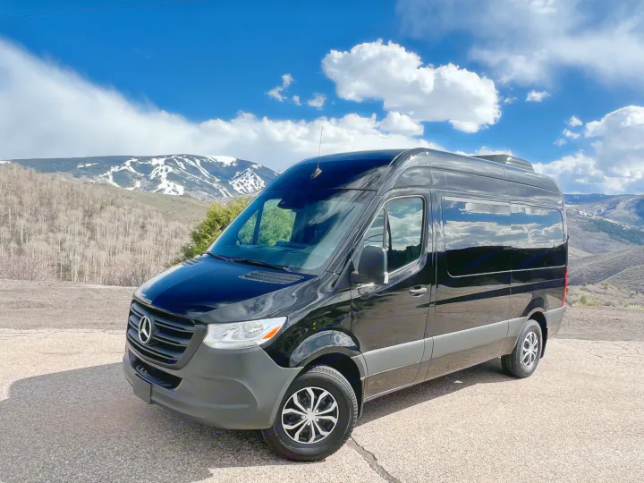 A black Mercedes Sprinter van, available for RV rental, is parked overlooking snowy mountains under a bright, cloudy sky.. Mercedes-Benz Sprinter 2023