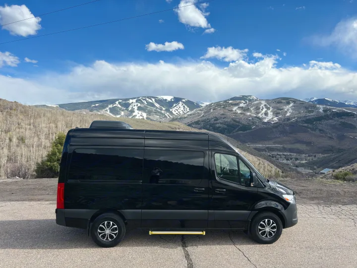 Black RV rental van parked on a paved overlook, with snow-capped mountains and a blue sky overhead.. Mercedes-Benz Sprinter 2023