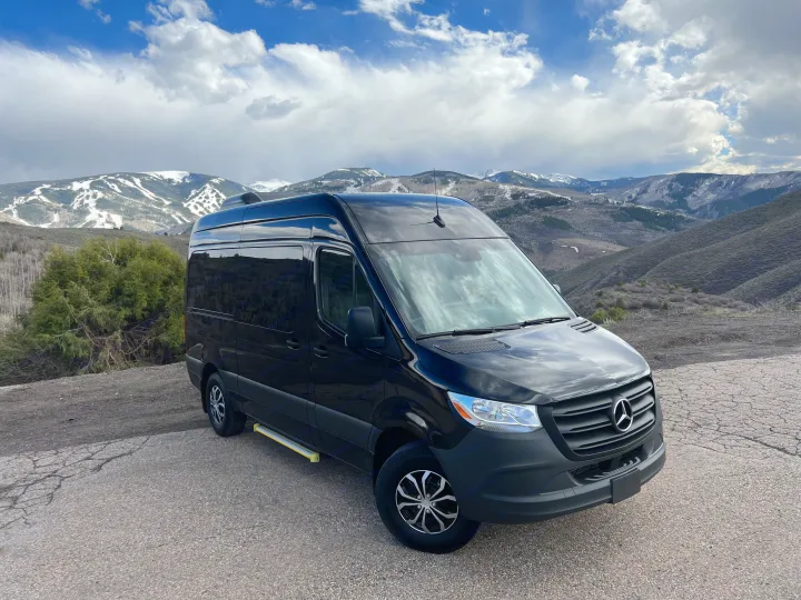Black Mercedes-Benz Sprinter RV rental van parked on a mountain overlook with snow-capped peaks and cloudy sky.. Mercedes-Benz Sprinter 2023