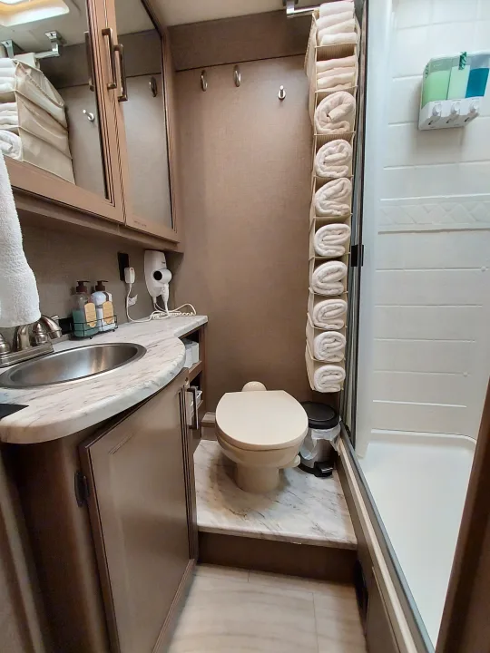 This RV rental bathroom features a compact toilet, sink, shower with dispensers, and neatly rolled towels in a hanging organizer.. Thor Coleman 2021