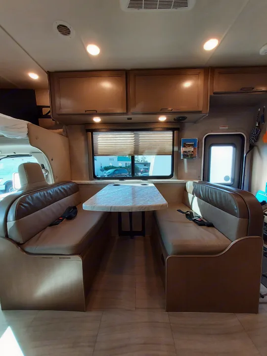 RV rental interior shows a bright dinette with a table, upholstered benches, overhead cabinets, and windows.. Thor Coleman 2021