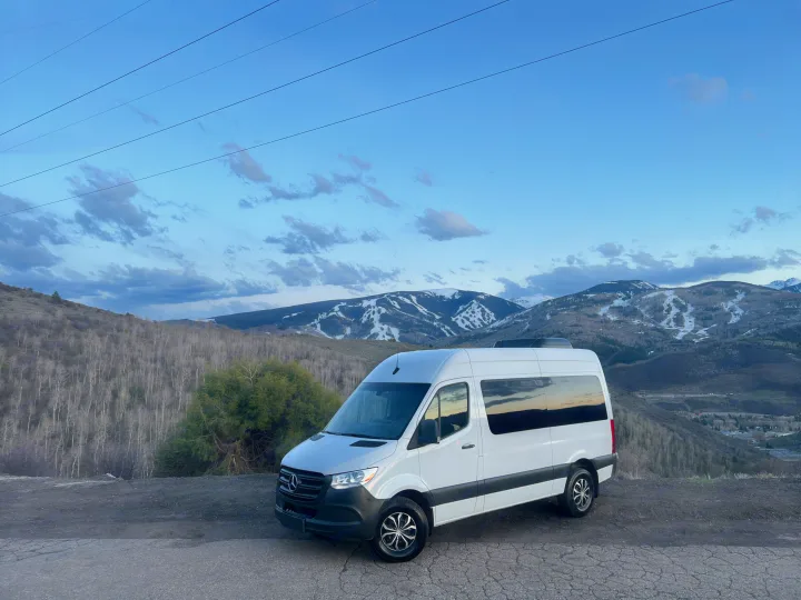 White Sprinter van RV rental parked on a mountain road with snowy peaks & blue sky. Perfect for scenic travel!. Mercedes-Benz Sprinter 2019