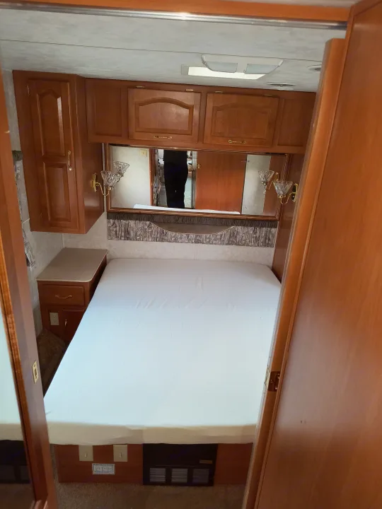 RV rental bedroom with a clean white bed, wooden overhead cabinets, a mirror, and a built-in nightstand.. Damon Corporation Daybreak 2001