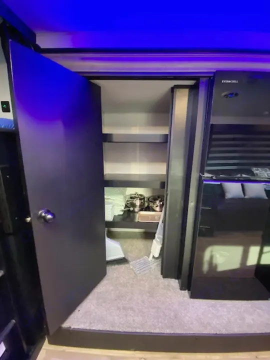 RV rental interior: Open pantry with shelves, pots, & cleaning tools on carpet. Blue lights illuminate the space next to a fridge and bedroom view.. Forest River Cherokee Grey Wolf 2022