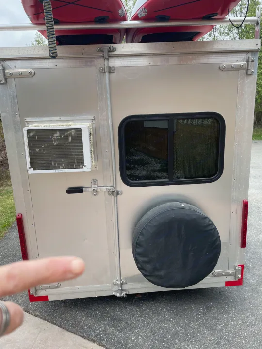 Rear view of a compact RV rental trailer, featuring kayaks overhead, a window AC unit, side window, and spare tire.. Weeroll Silver cloud 2020