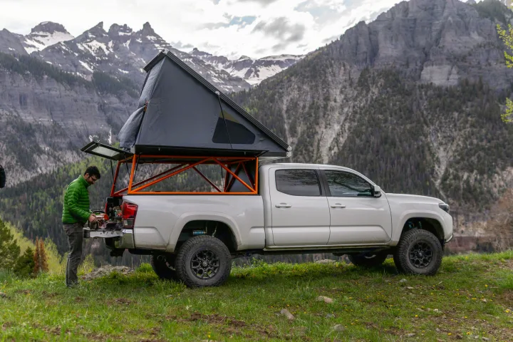 White truck with rooftop tent on a mountain pass. Man preps meal. This setup is great for an RV rental adventure.. Other Other 2019