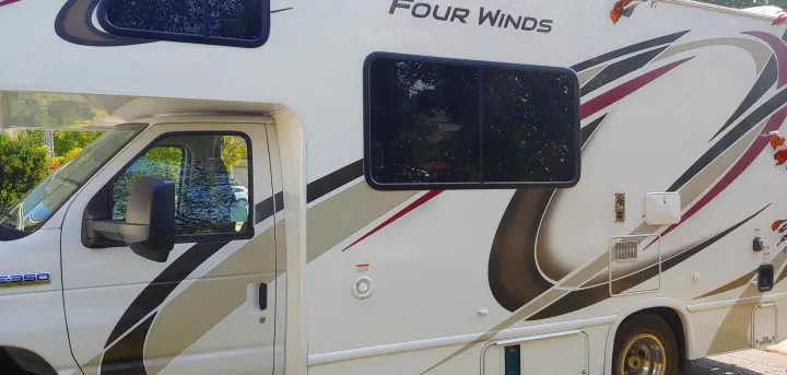 Close-up side view of a white Four Winds E-350 motorhome. This RV rental shows tinted windows & decorative decals.. Thor Four Winds 2020