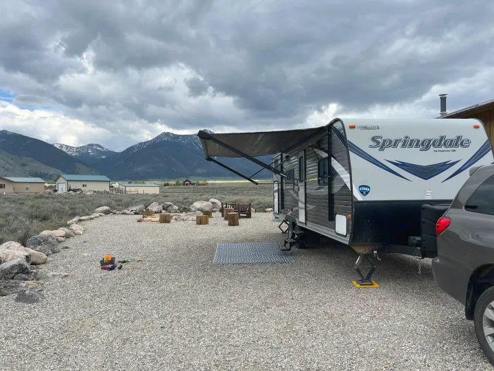 Springdale RV rental parked on a gravel site with stunning mountain views, fire pit ready for camping under cloudy skies.. Keystone RV Springdale 2019