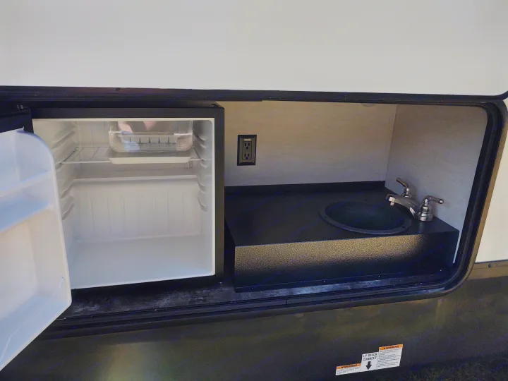 RV rental outdoor kitchen setup with an open mini-fridge, sink, and electrical outlet.. Forest River Sabre 2021