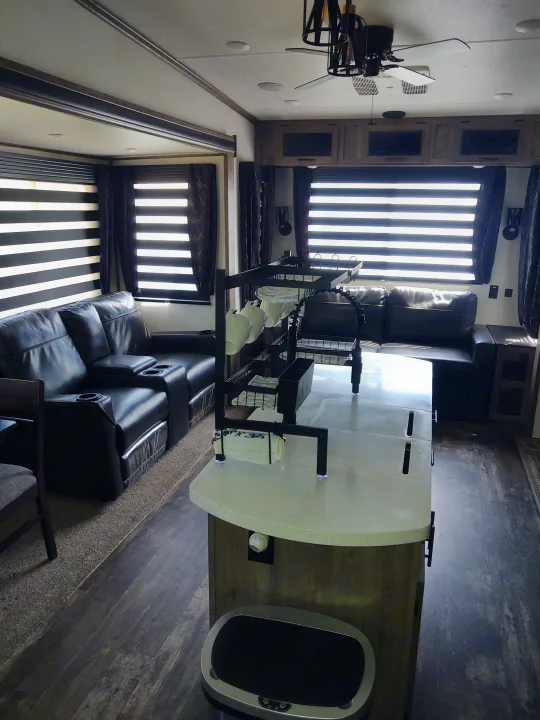 View of a modern RV rental interior, showing a spacious kitchen island, black living room recliners, and bright windows.. Forest River Sabre 2021