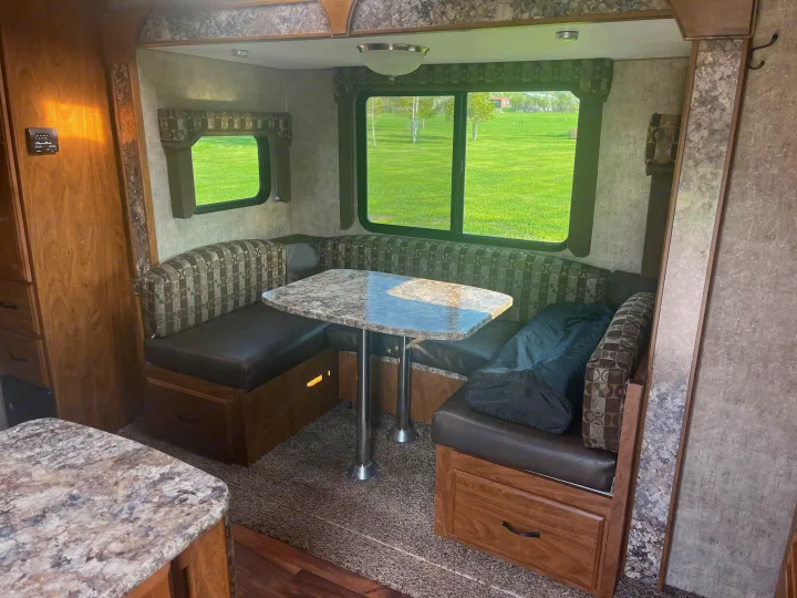 Bright RV rental interior features a U-shaped dinette with a granite-look table and large windows overlooking a green field.. Outdoors RV Creekside 2017