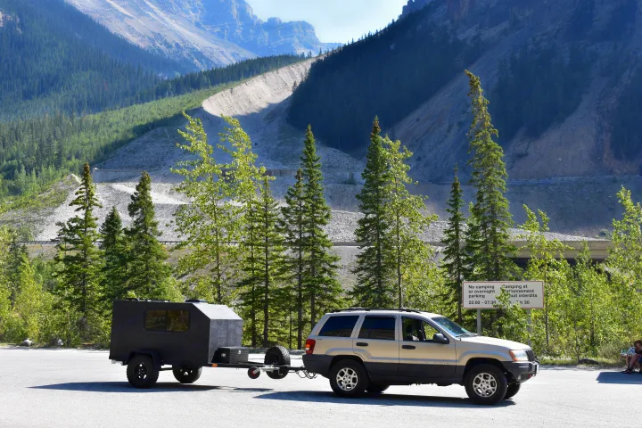 Tan SUV towing a dark camper trailer (possible RV rental) in a scenic mountain parking lot, next to a "no overnight parking" sign.. U-Bilt U-Bilt 2021