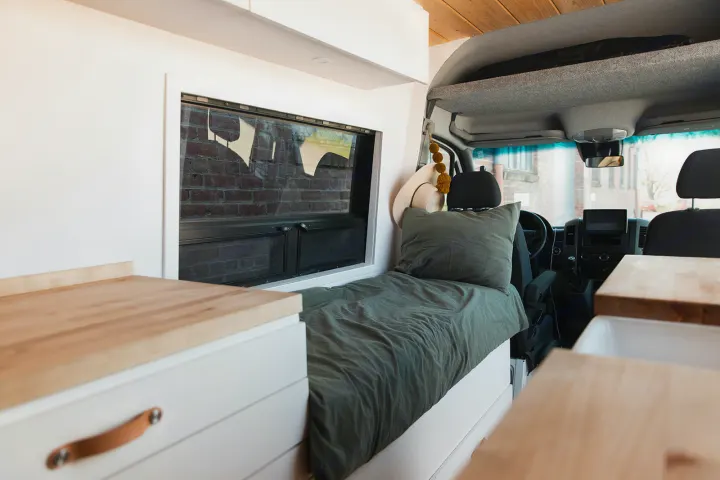 Cozy RV rental interior featuring a bed, window, and wood countertops, with the driver's cab visible.. Mercedes-Benz Sprinter 2014