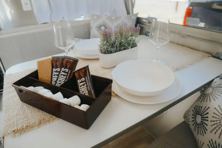 Cozy RV rental dining setup: S'mores ingredients, white dishes, wine glasses, and a lavender plant on a patterned mat.. Airstream Bambi 2023