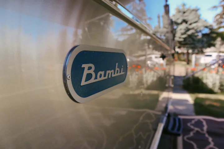 Close-up of the "Bambi" emblem on a polished Airstream travel trailer, reflecting outdoor scenery. Consider an RV rental!. Airstream Bambi 2023