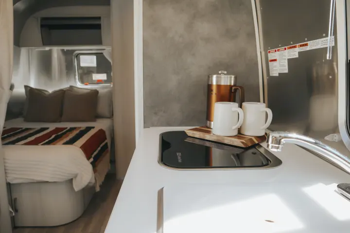 Cozy RV rental interior featuring a modern kitchen area with a Stanley coffee press & mugs, opposite a comfortable bed with striped blankets.. Airstream Bambi 2023