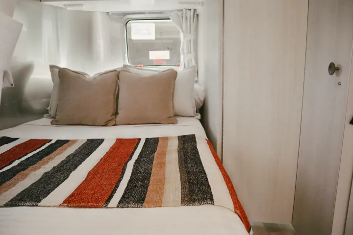 Cozy bed inside a clean, modern RV rental, with a colorful striped blanket and pillows. Ideal for comfortable travel.. Airstream Bambi 2023