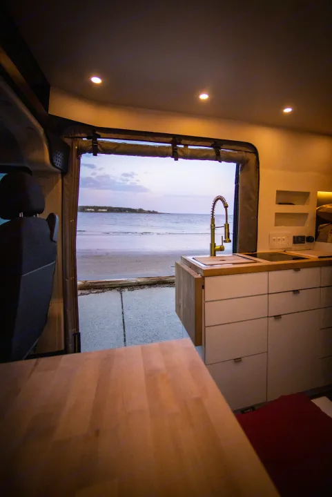 RV rental kitchen view: Modern van interior with golden faucet, ocean sunset through open door.. Dodge Ram Promaster 3500 2021