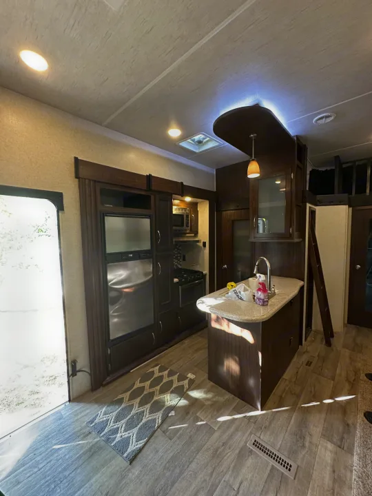 Modern RV rental kitchen & living space: island sink, full fridge, stove, and dark wood cabinetry with light flooring.. Heartland Torque 2018