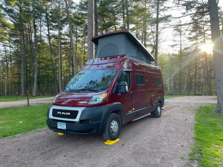 This RV rental in a serene wooded campsite features a red pop-top camper van ready for adventure.. Winnebago Solis 2021