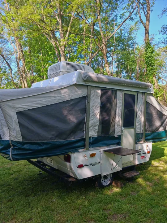 This white and green pop-up camper RV rental, with an AC unit, is set up on a grassy campsite under lush trees.. Coleman Other 2002