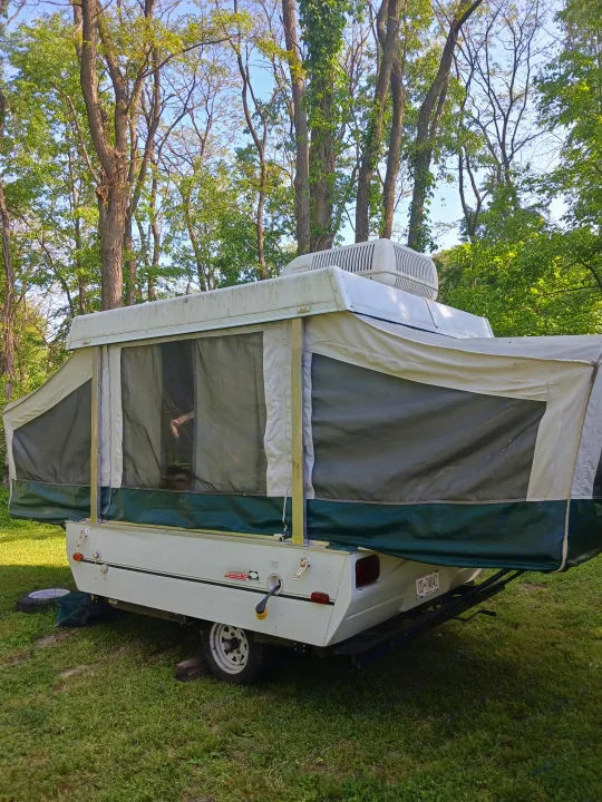 This pop-up camper, ideal for an RV rental, is set up in a wooded campsite, ready for outdoor travel and camping.. Coleman Other 2002
