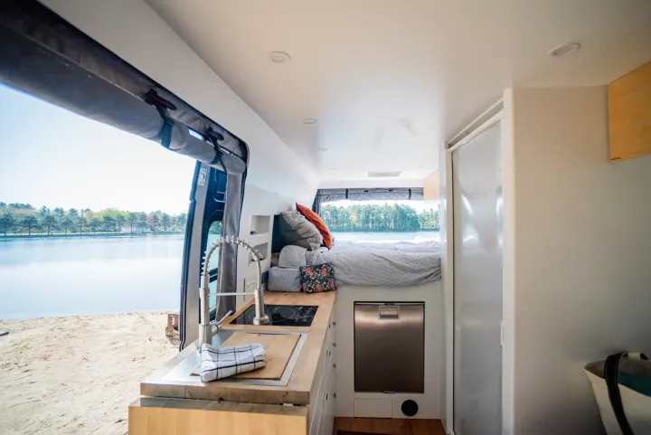 This RV rental showcases a modern interior with a kitchen, cozy bed, and a stunning lake view from the open doors.. Dodge Ram Promaster 3500 2021
