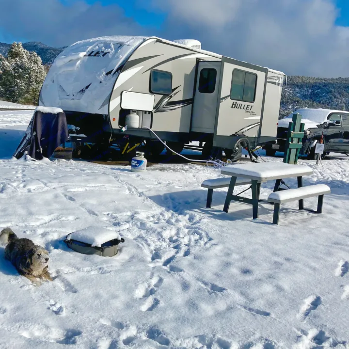 A snow-covered RV rental sits in a wintry campsite, a fluffy dog by a snowy picnic table, mountains in the distance.. Keystone RV Bullet Ultra Lite 2020