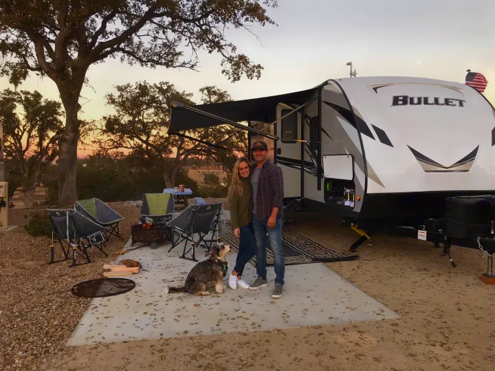 A couple and their dog pose in front of a white Bullet RV rental at a scenic campsite with a fire pit.. Keystone RV Bullet Ultra Lite 2020