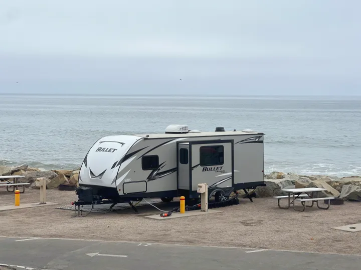 Oceanfront rv rental travel trailer at a coastal campsite, next to the sea and rocks under a cloudy sky. Picnic tables visible.. Keystone RV Bullet Ultra Lite 2020