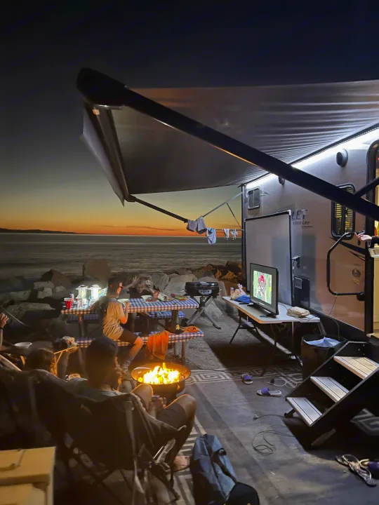 Evening by the water with an RV rental: Friends gather around a campfire, watching TV under the awning at sunset.. Keystone RV Bullet Ultra Lite 2020
