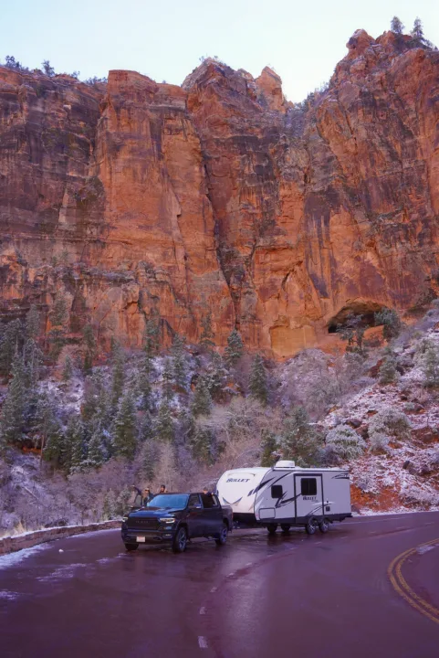 Winter wonderland rv rental adventure: a truck towing a travel trailer pauses on a wet road beside towering snowy red rock cliffs.. Keystone RV Bullet Ultra Lite 2020