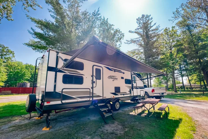 Sunny day at an RV rental campsite: A white travel trailer with an awning, picnic table, and truck surrounded by tall trees.. Grand Design Reflection 2021